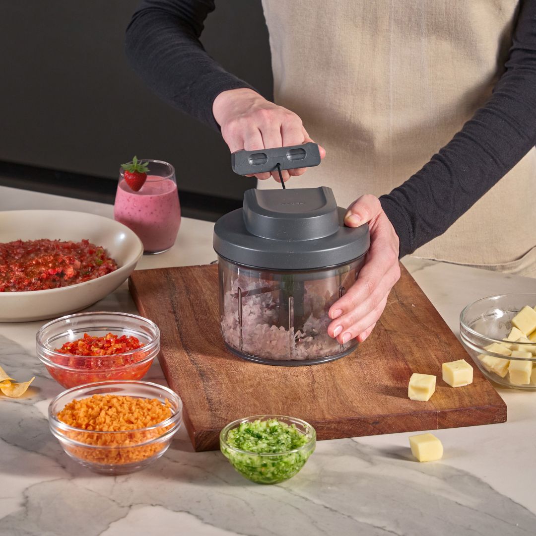 A manual food chopper filled with chopped vegetables sits on a kitchen counter, with whole vegetables and a cutting board in the background.