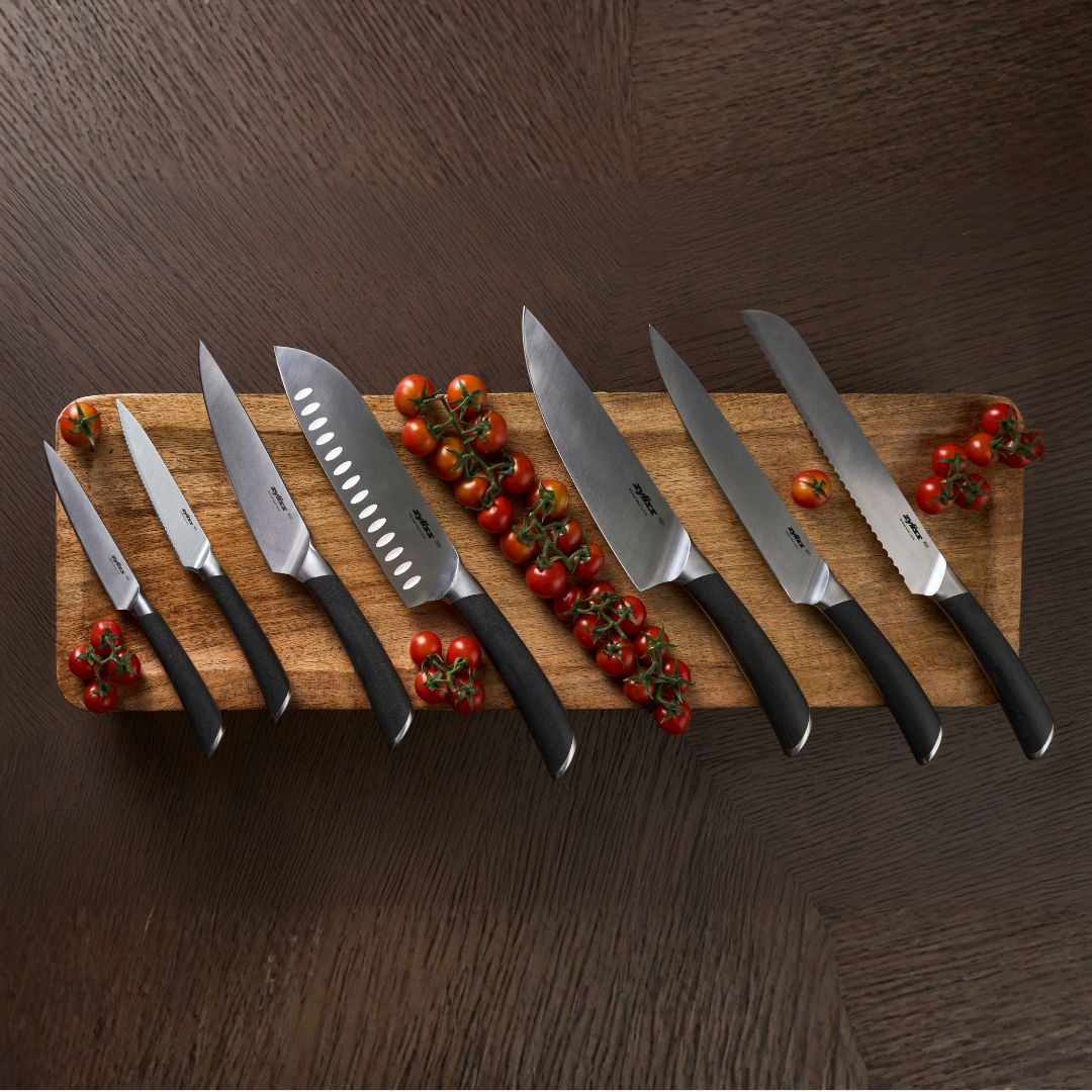 A bowl of vegetable stew sits on a wooden board with two slices of bread, three kitchen knives, and a potted basil plant on a gray surface.