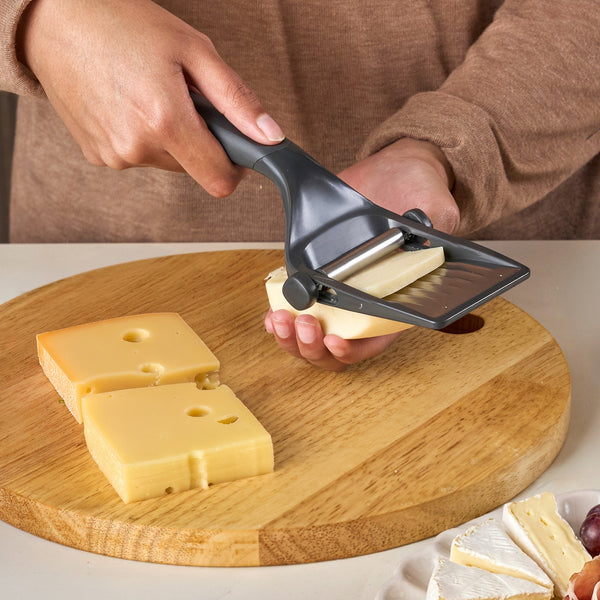 A person uses the Zyliss Utility Cheese Plane, featuring an adjustable stainless steel blade, to cut cheese slices from a block on a round wooden cutting board.