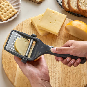 Using the Zyliss Utility Cheese Plane with a stainless steel blade, a person cuts cheese on a wooden board. Nearby, sliced cheese, crackers, and bread are visible.