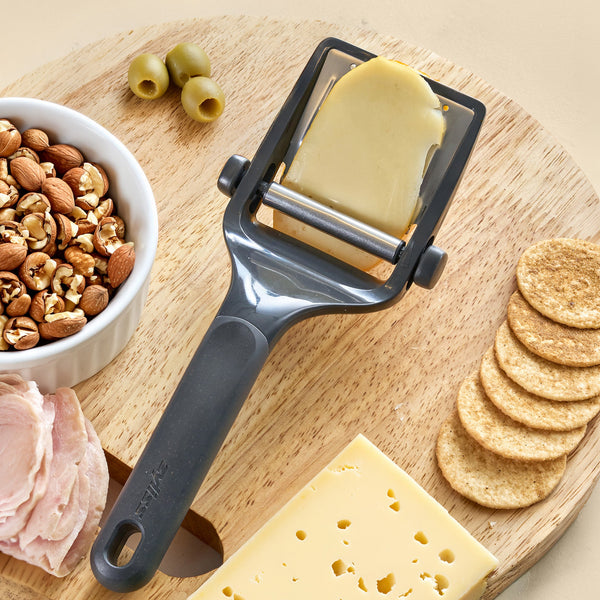 The Zyliss Utility Cheese Plane sits on a wooden board with a cheese slice, alongside a cheese block, round crackers, sliced deli meat, mixed nuts in a bowl, and green olives.