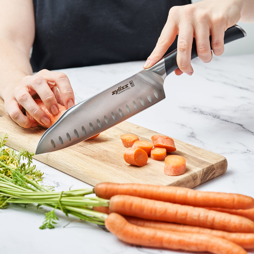 The Zyliss Comfort Pro Santoku Knife 18cm/7 features German stainless steel, a black ergonomic handle, and oval blade indentations, displayed against a light background. 