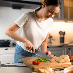 A woman in a white t-shirt smiles as she chops green herbs with the Zyliss Chef Knife 15cm/6, featuring an ergonomic handle, on a wooden cutting board in a modern kitchen. Cheese and tomatoes are also on the board. 