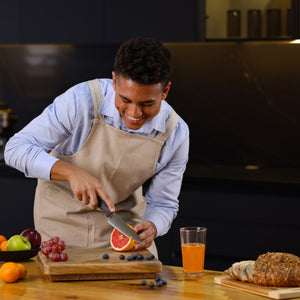 A smiling person in an apron uses the Zyliss Chef Knife 15cm / 6 with an ergonomic handle to slice a grapefruit on a wooden cutting board in a sustainable kitchen, surrounded by grapes, apples, blueberries, juice, and sliced bread. 