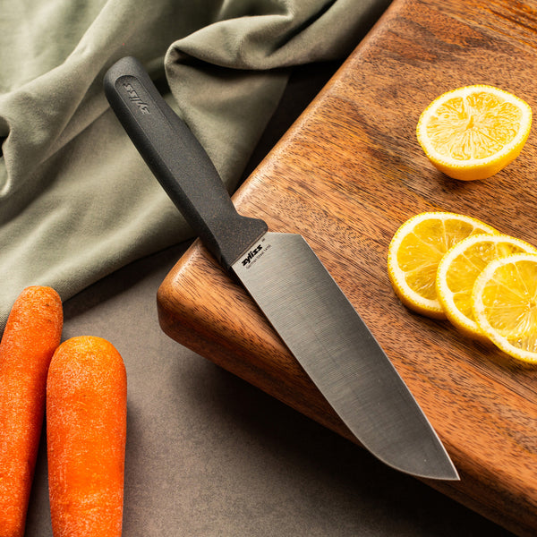 A Zyliss Santoku Knife 15cm / 6 with an ergonomic handle lies on a wooden cutting board beside sliced lemons, whole carrots, and a green cloth—all arranged on a gray surface for an inviting kitchen scene. 