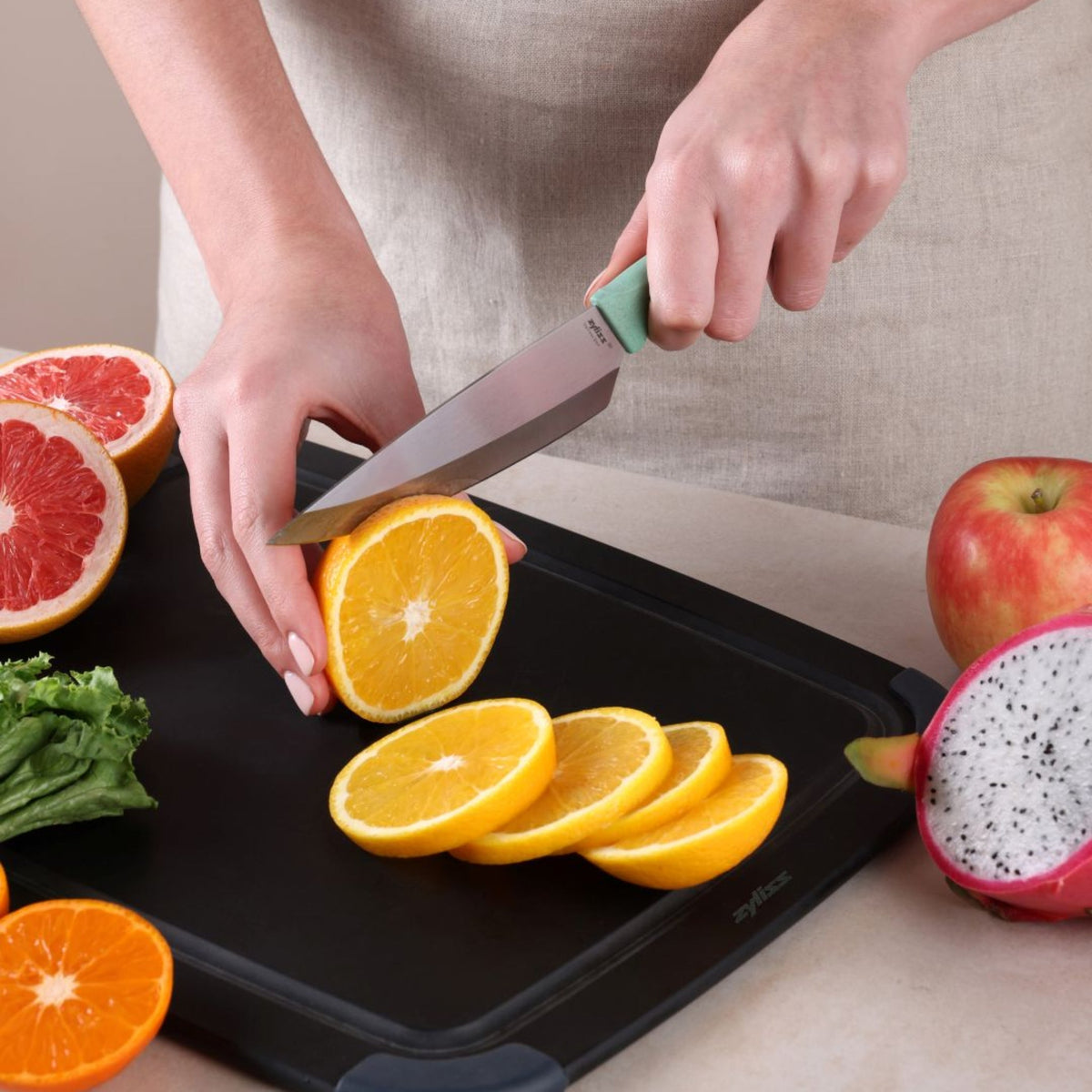 A person slices an orange with the Zyliss Classic Utility Knife on a black cutting board surrounded by lettuce, grapefruit, apple, and dragon fruit.