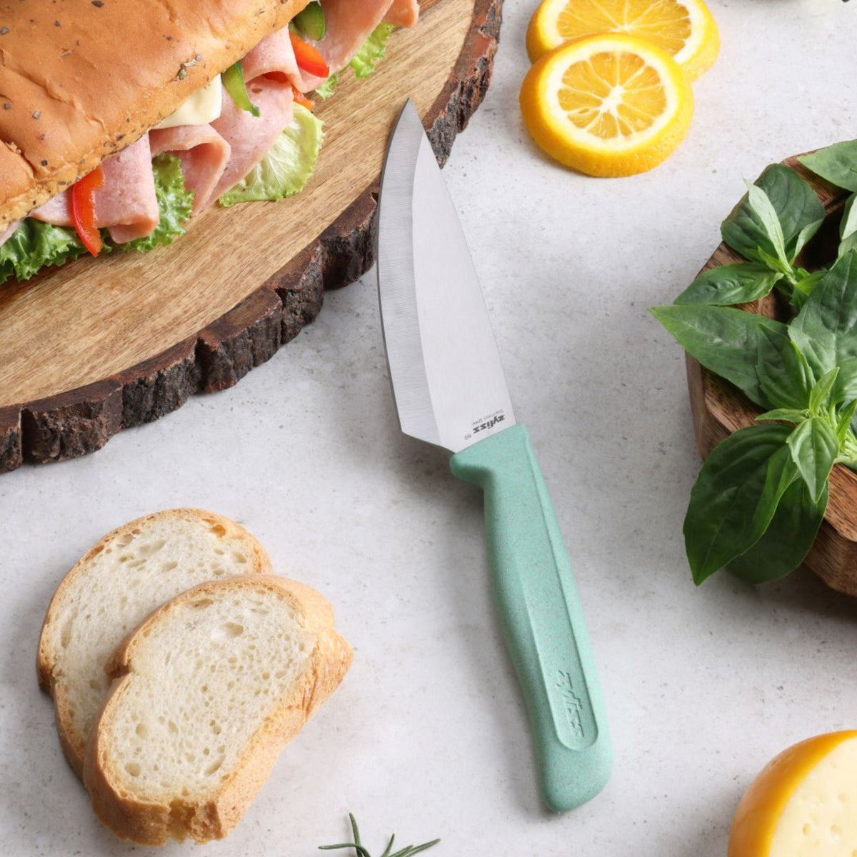 A Zyliss Classic Utility Knife with a mint green handle rests on a table beside two slices of bread, a sandwich, lemon slices, fresh basil, and a wooden bowl.