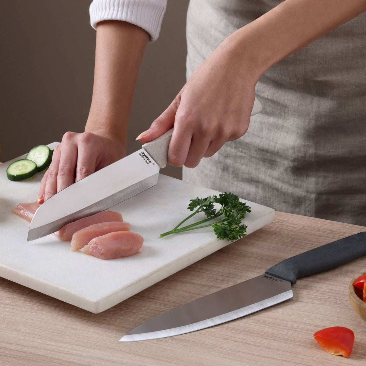 A person slices raw chicken on a white cutting board using a sharp stainless steel chef knife from the Zyliss Classic 2 Piece Chef & Santoku Knife Set. Cucumber slices, parsley, a tomato piece, and the santoku knife from the same set are also on the board.