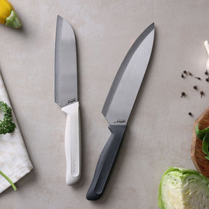 The Zyliss Classic 2 Piece Chef & Santoku Knife Set—one knife with a white handle and the other with a black—rests on a light countertop near cabbage, parsley, yellow squash, scattered peppercorns, and a towel.