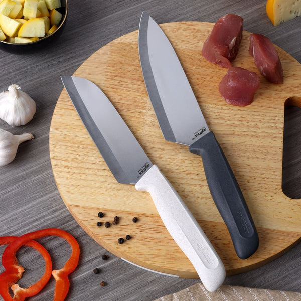The Zyliss Classic 2 Piece Chef & Santoku Knife Set, with white and black handles, displayed on a round wooden cutting board surrounded by raw meat, garlic, cheese, black peppercorns, pepper slices, and freshly chopped vegetables.
