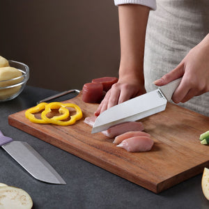 A person slices raw chicken on a wooden cutting board with a knife from the Zyliss Classic 2 Piece Santoku Knife Set, with yellow bell pepper and potatoes on the countertop.