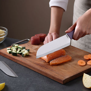 Using the Zyliss Classic 2 Piece Santoku Knife Set, a person slices raw salmon on a wooden cutting board surrounded by chopped zucchini, carrots, and tuna, while the second knife rests on the counter.