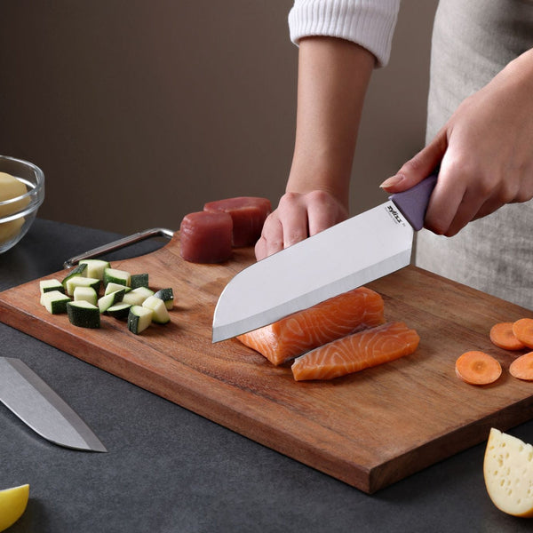 Using the Zyliss Classic 2 Piece Santoku Knife Set, a person slices raw salmon on a wooden cutting board surrounded by chopped zucchini, carrots, and tuna, while the second knife rests on the counter.