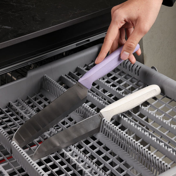 A hand places a knife with a lavender handle into a dishwasher rack next to a similar knife with a white handle, both from the Zyliss Classic 2 Piece Santoku Knife Set.
