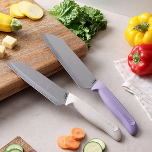 Two knives from the Zyliss Classic 2 Piece Santoku Knife Set, with white and purple ergonomic handles, rest on a counter near sliced vegetables, a cutting board, lettuce, yellow squash, and red and yellow bell peppers.