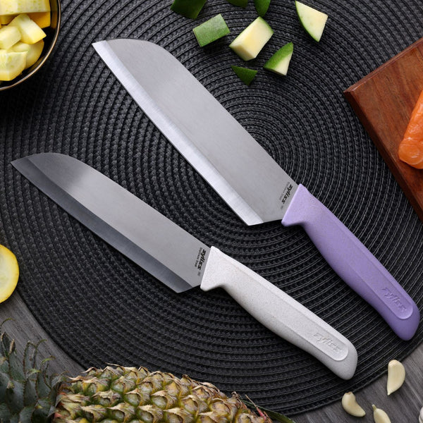 Two knives from the Zyliss Classic 2 Piece Santoku Knife Set with white and lavender handles displayed on a black textured mat alongside cut vegetables, fresh pineapple, and a wooden board topped with salmon.