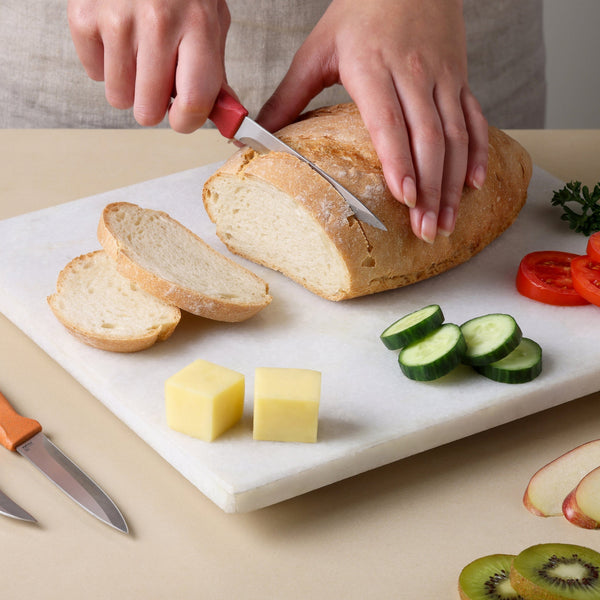 A person slices a loaf of bread on a cutting board with a knife from the Zyliss Classic 3 Piece Peeling & Paring Knife Set, with sliced cucumber, tomato, cheese cubes, and fruit slices arranged next to the other knives.