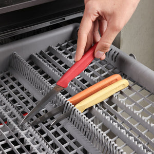 A hand places a red-handled knife from the Zyliss Classic 3 Piece Peeling & Paring Knife Set into a dishwasher utensil rack, where the orange- and yellow-handled knives are already placed.