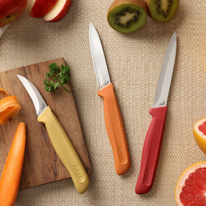 Three stainless steel knives from the Zyliss Classic 3 Piece Peeling & Paring Knife Set with yellow, orange, and red handles arranged on a woven surface next to sliced kiwi, grapefruit, apple, and a wooden board with parsley.