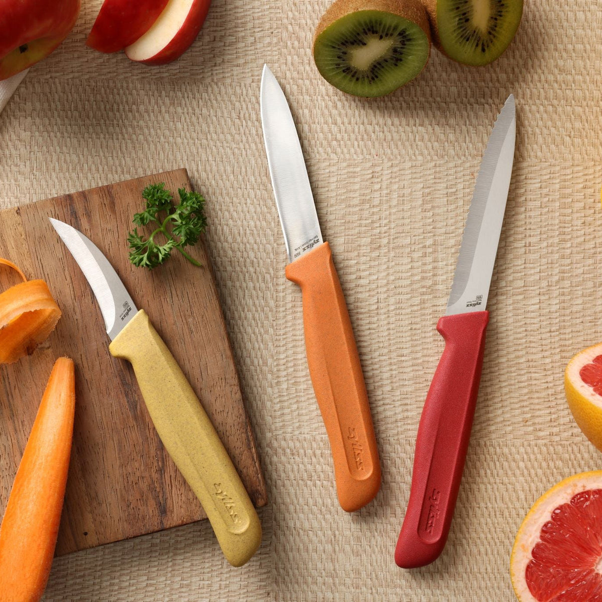 Three stainless steel knives from the Zyliss Classic 3 Piece Peeling & Paring Knife Set with yellow, orange, and red handles arranged on a woven surface next to sliced kiwi, grapefruit, apple, and a wooden board with parsley.