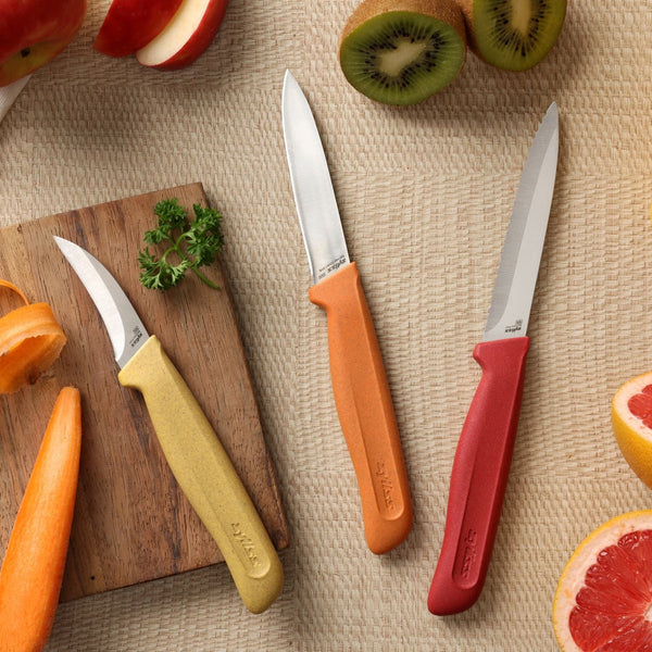 Three stainless steel knives from the Zyliss Classic 3 Piece Peeling & Paring Knife Set with yellow, orange, and red handles arranged on a woven surface next to sliced kiwi, grapefruit, apple, and a wooden board with parsley.