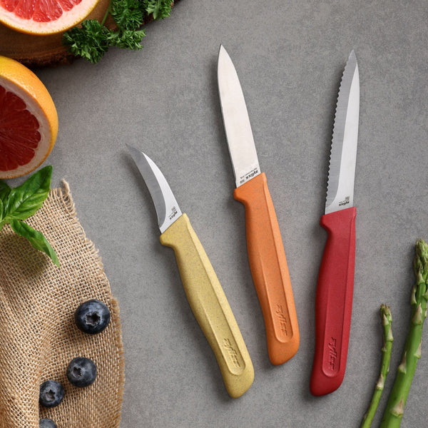 Three knives from the Zyliss Classic 3 Piece Peeling & Paring Knife Set with yellow, orange, and red handles on a gray surface surrounded by blueberries, grapefruit, basil, parsley, asparagus, and burlap fabric.