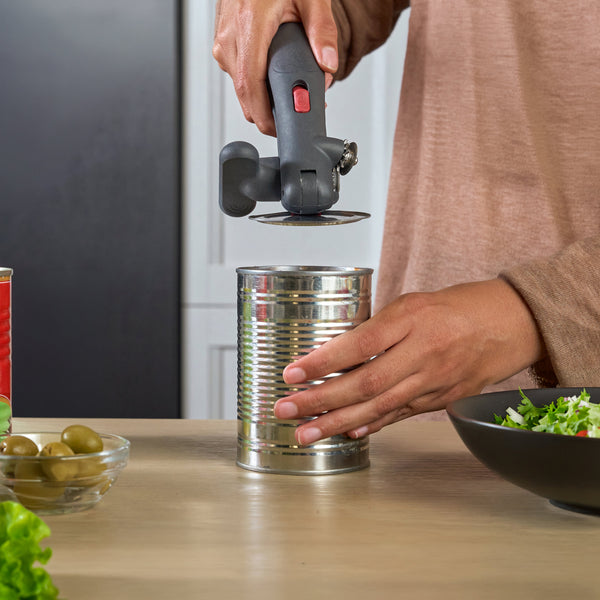 A person using a cordless angle grinder can opener to cut open a metal can on a kitchen counter, with canned olives, salad, and other kitchen items nearby.