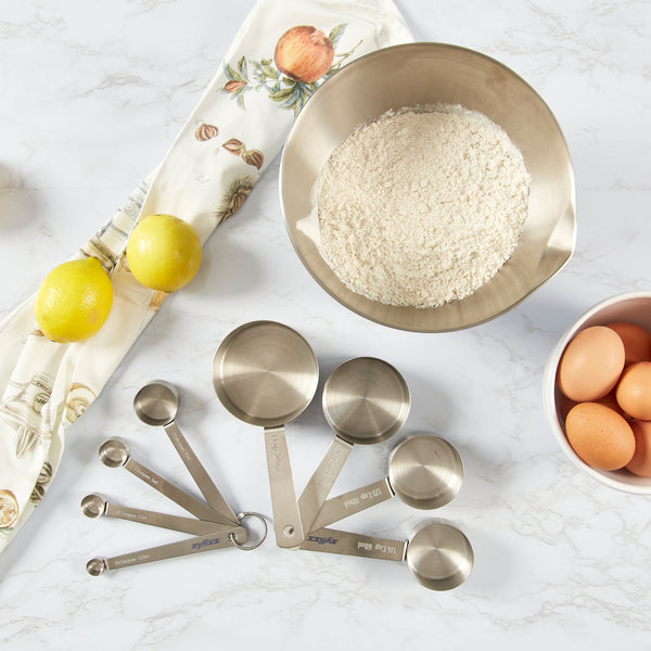 A bowl of flour, a bowl with four eggs, two lemons, Zyliss Stainless Steel Measuring Spoons, and a decorative kitchen towel are arranged on a marble countertop—perfect for any baking and cooking enthusiast.