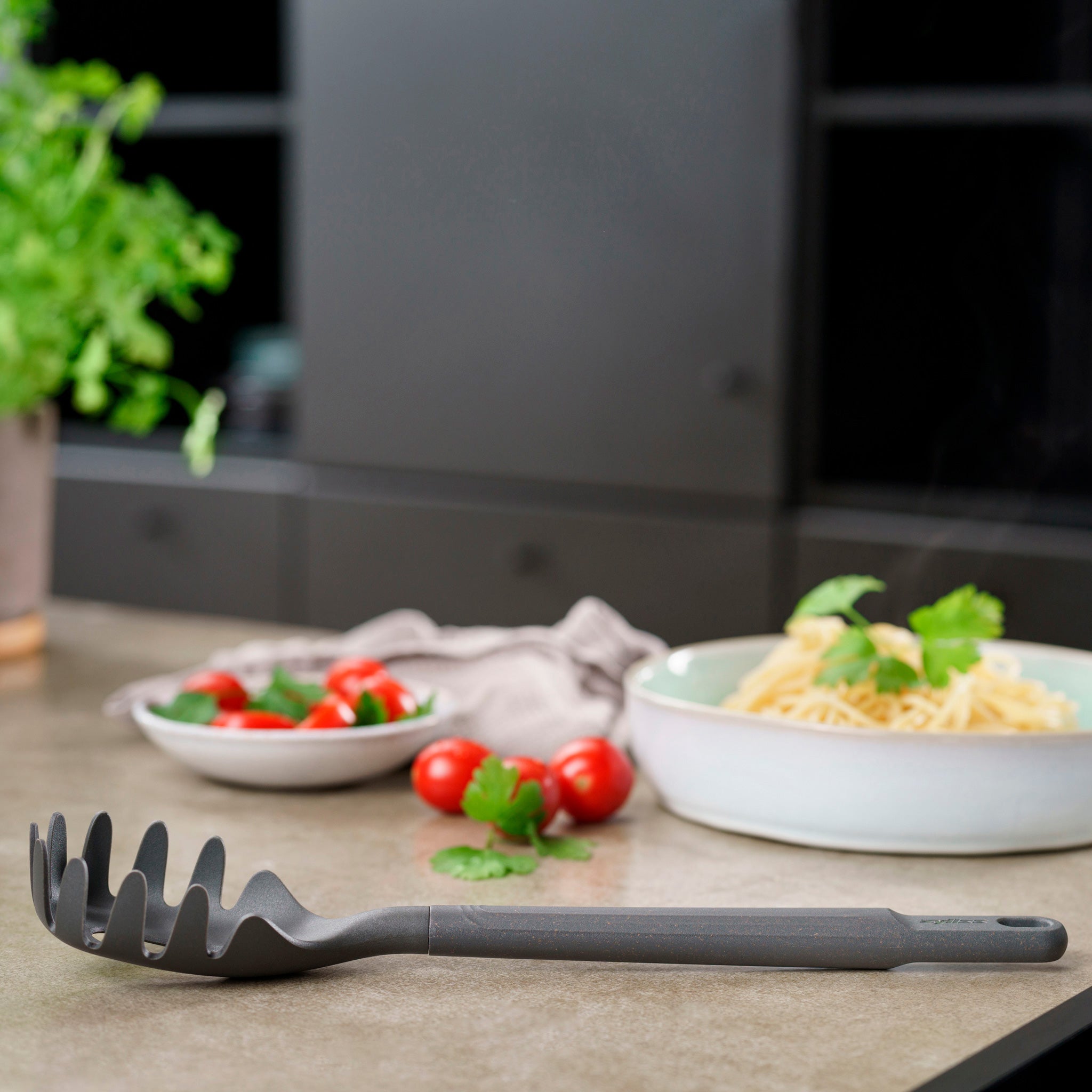 A Zyliss Spaghetti Server, heat-resistant and black, sits on a kitchen counter with a bowl of pasta, a plate of cherry tomatoes, fresh cilantro, and a potted plant in the background.