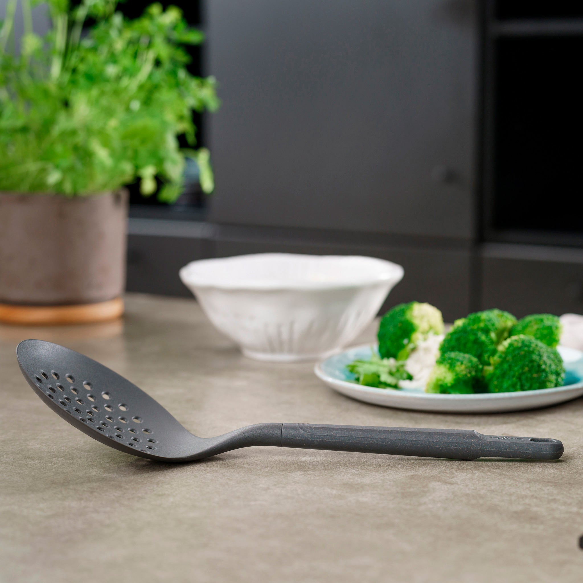 A Zyliss Skimmer with an ergonomic handle sits on a kitchen counter next to a small plate of broccoli and cauliflower, a white bowl, and a potted plant in the background.