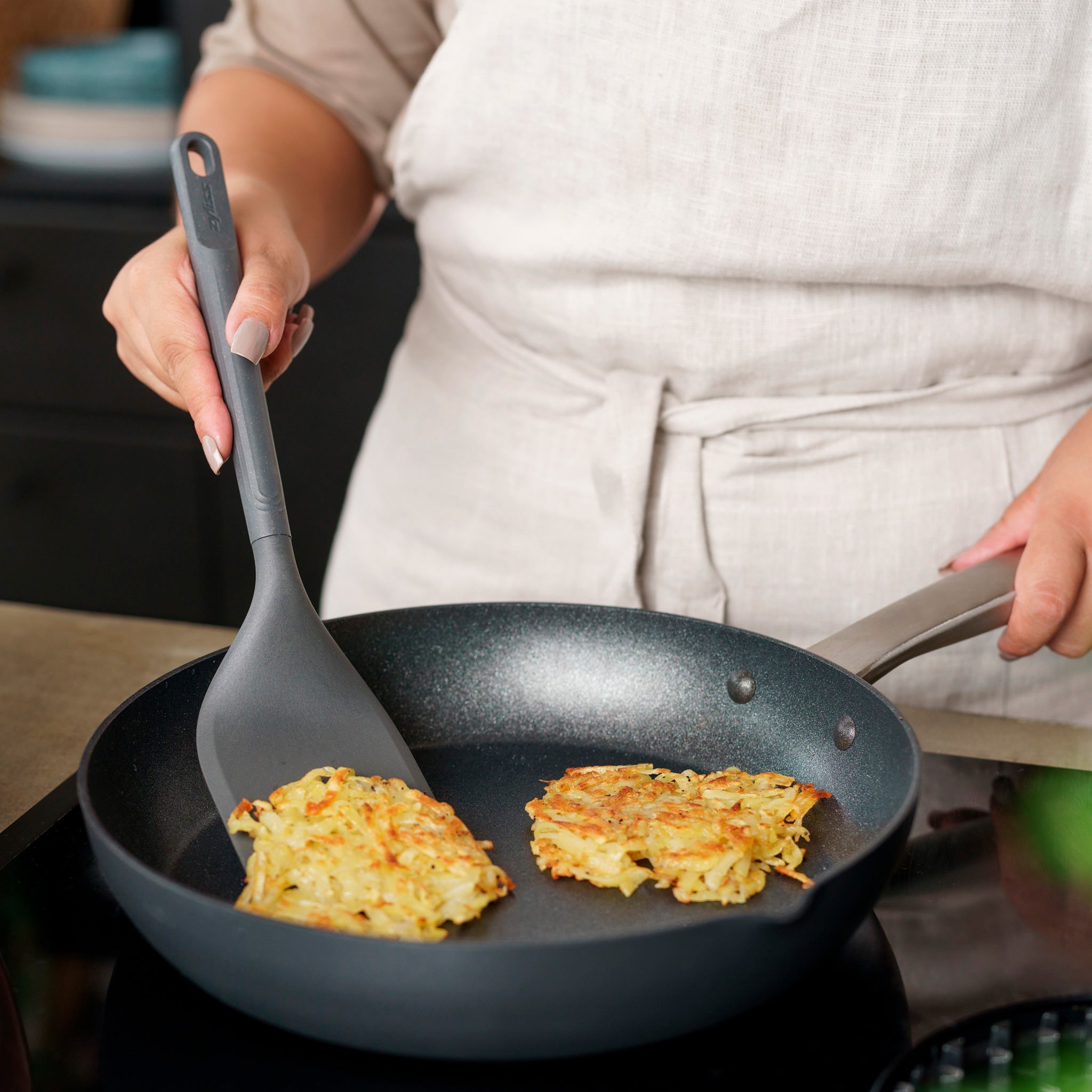 Wearing a beige apron, a person uses the Zyliss Angled Turner to cook two potato pancakes in a nonstick frying pan on the stovetop.