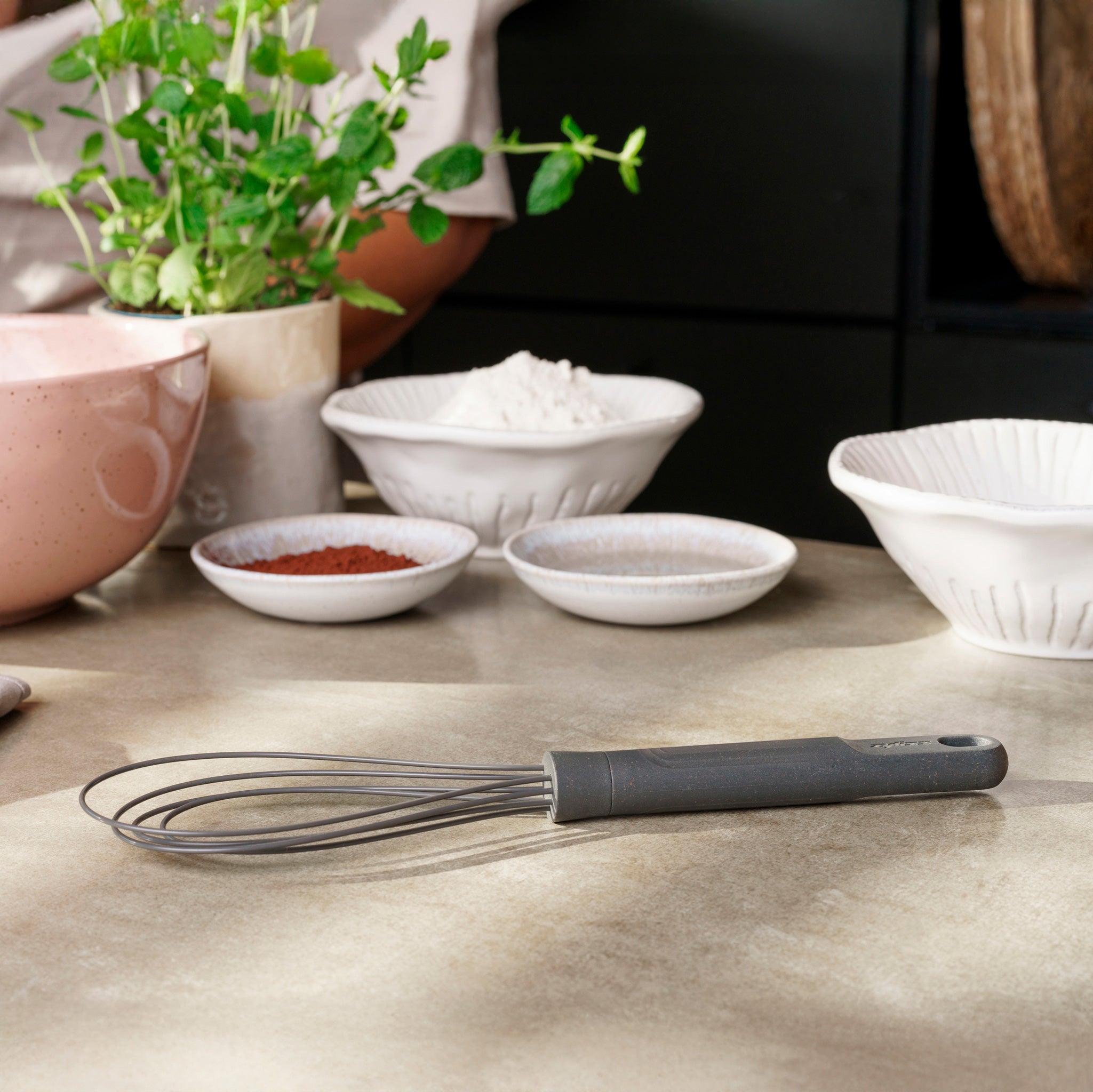 The Zyliss Flat Whisk Silicone with a gray handle rests on a countertop alongside ceramic bowls of baking ingredients and a pink mixing bowl, while fresh green herbs provide a lively backdrop.