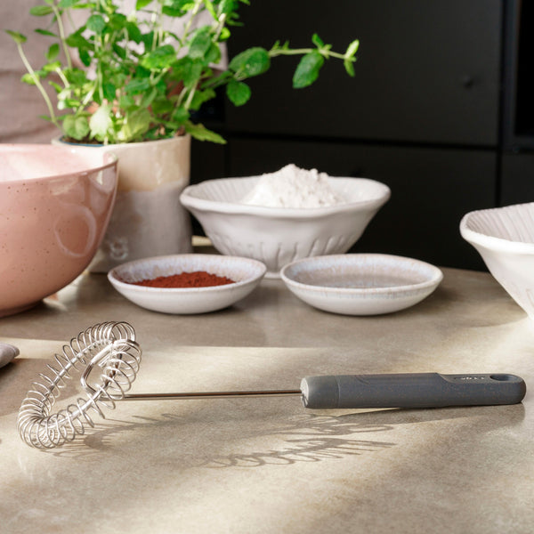 A Zyliss Gravy Whisk rests on a kitchen countertop with bowls of cocoa powder, flour, and sugar for sauce prep; a pink mixing bowl and a green potted herb are in the background. 