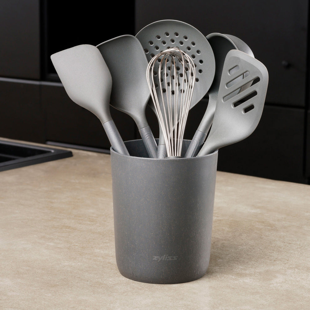 The Zyliss 7-Piece Utensil Set includes black kitchen tools—whisk, spatulas, spoon, and slotted spoon—organized in a matching container. Each features ergonomic handles and heat-resistant PBT for safe, comfortable use. 