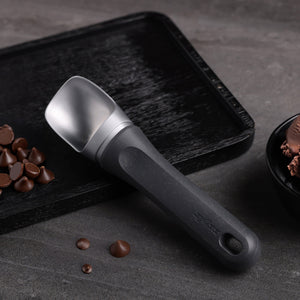 A Zyliss Ice Cream Scoop with an eco-friendly handle sits on a textured black tray beside chocolate chips and a bowl of chocolate ice cream, arranged on a gray surface. 