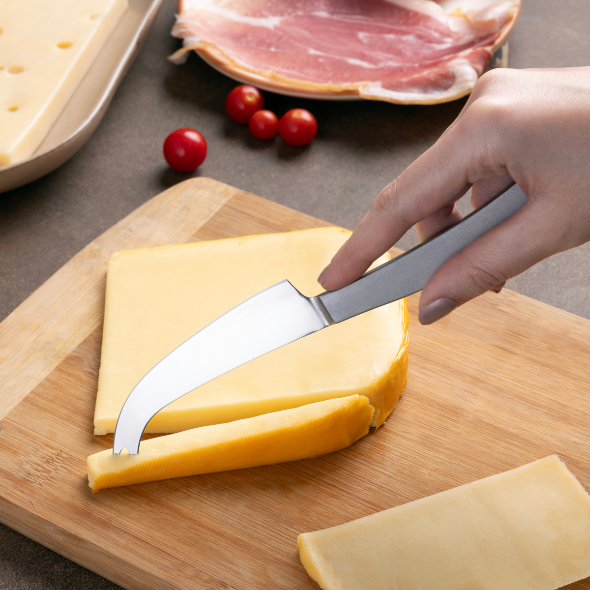 A hand slices a wedge of yellow cheese on a wooden cheese board using the Zyliss Signature Universal Cheese Knife, with sliced cheese, ham, cherry tomatoes, and Swiss cheese in the background.
