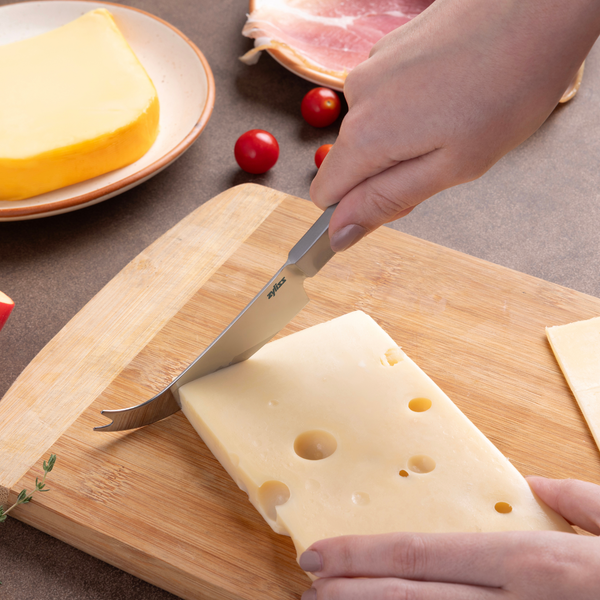 Using the Zyliss Signature Universal Cheese Knife, a person slices Swiss cheese on a wooden cutting board next to a wedge of yellow cheese, slices of meat, and cherry tomatoes.