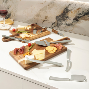The Zyliss Signature 3 Piece Cheese Knife & Acacia Serving Board Set displays assorted cheeses, crackers, and meats on a white marble countertop, with a glass of red wine and napkin in the background.
